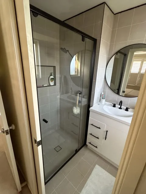 Black-framed shower screen beside a white vanity and round bathroom mirror