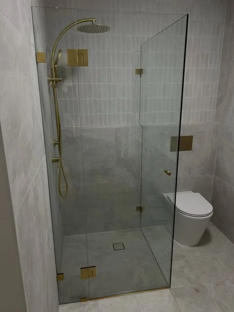 Frameless glass shower screen with brass hinges and handle in a grey tiled bathroom