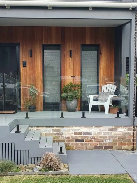 Glass balustrade with black spigots across a timber-clad front veranda above brick steps