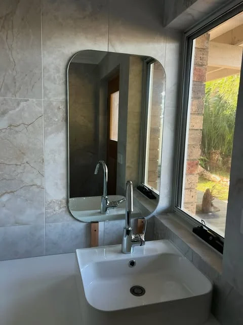 Custom rounded rectangle bathroom mirror above a white basin beside a large window